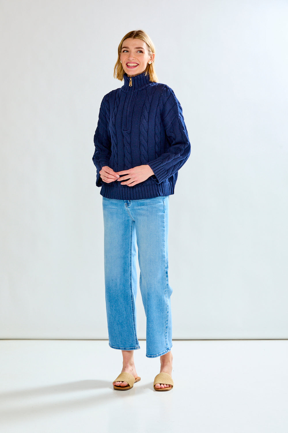 Woman wearing a navy cable knit sweater and blue jeans on a white background