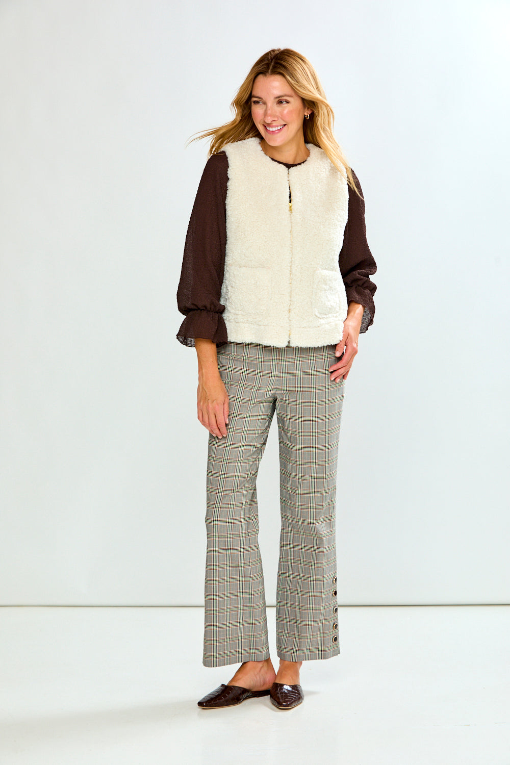 A woman modeling a brown long-sleeve top with a cream sherpa vest, paired with green and white plaid trousers, against a light gray studio background.