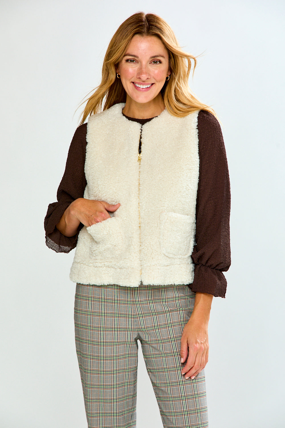 A woman modeling a brown long-sleeve top with a cream sherpa vest, paired with green and white plaid trousers, against a light gray studio background.
