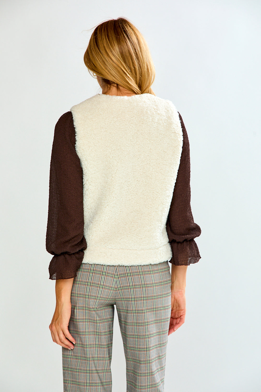 A woman modeling a brown long-sleeve top with a cream sherpa vest, paired with green and white plaid trousers, against a light gray studio background.