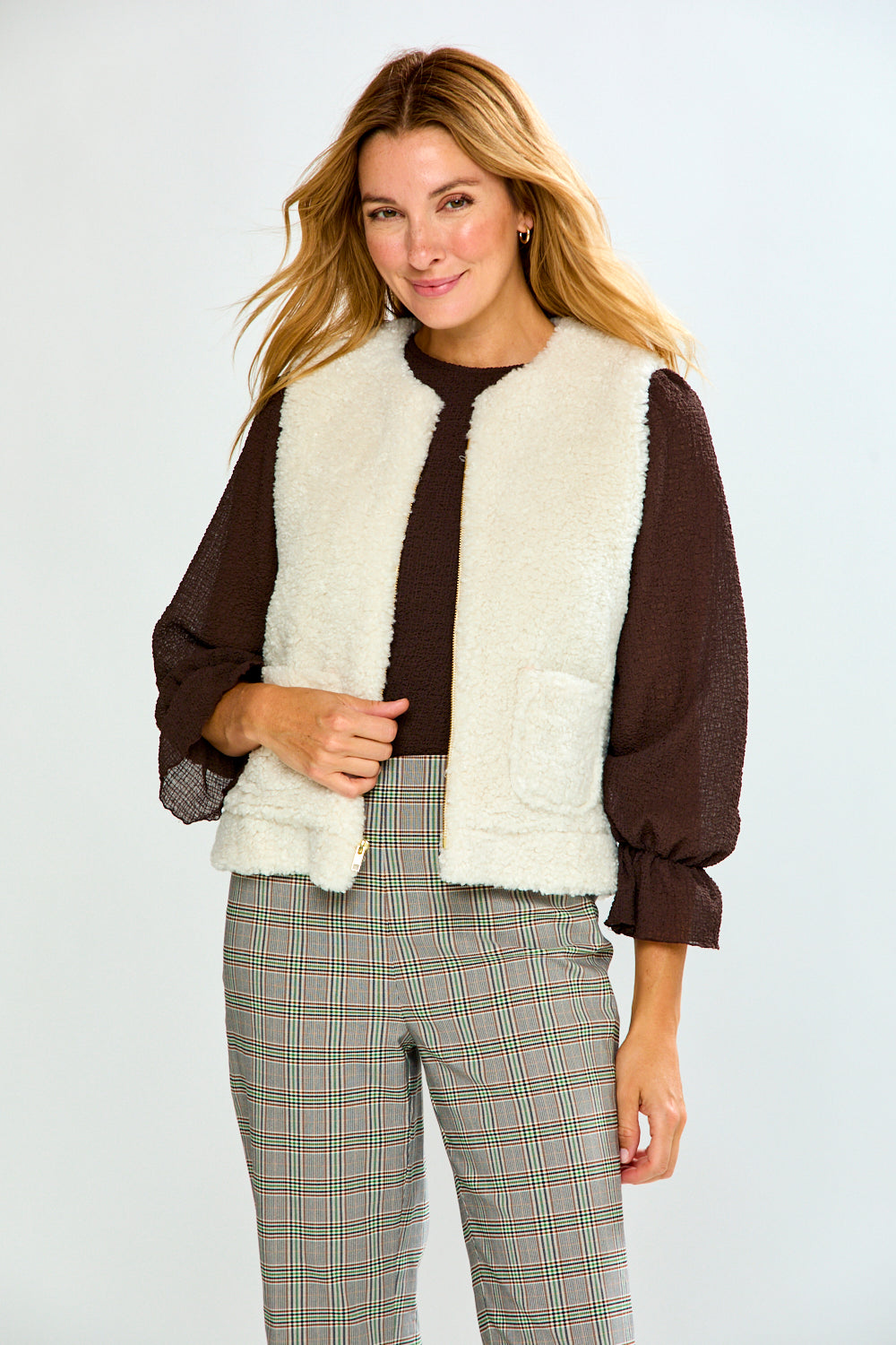 A woman modeling a brown long-sleeve top with a cream sherpa vest, paired with green and white plaid trousers, against a light gray studio background.