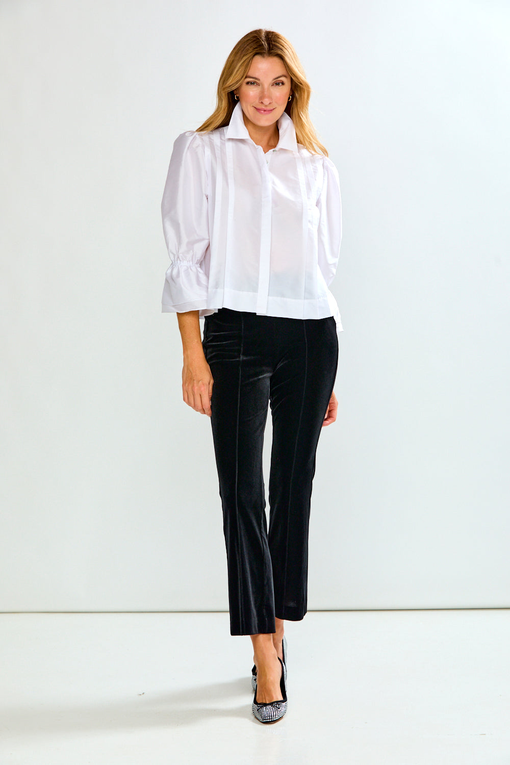 A woman modeling a white long-sleeve blouse with ruffled cuffs and black cropped trousers, standing against a plain white studio background.
