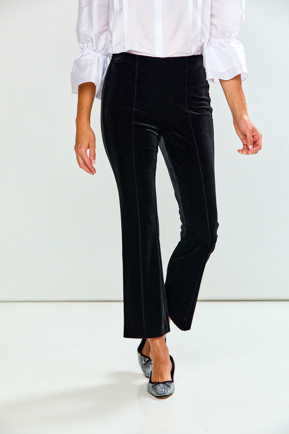 A model wearing black velvet high-waisted cropped flare pants, paired with a white blouse and black pointed flats, shown from the waist down against a white studio background.