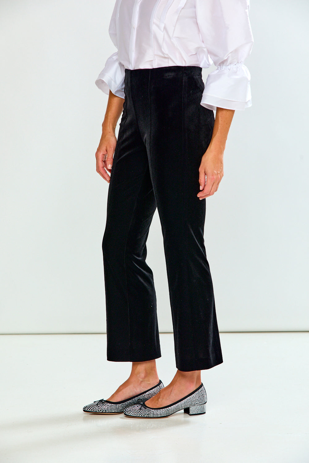 A model is shown from the waist down wearing black cropped velvet trousers, a white blouse with ruffled sleeves, and sparkly silver flats, set against a plain white studio background.