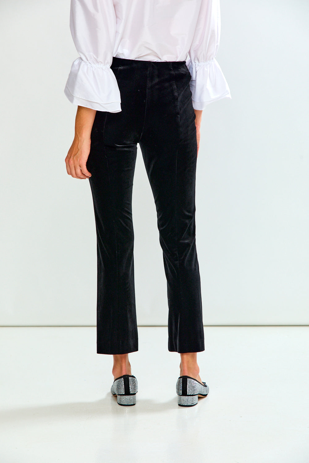 A model from the back wearing black velvet cropped trousers, a white blouse with ruffled sleeves, and black-and-white patterned shoes, set against a clean white studio background.