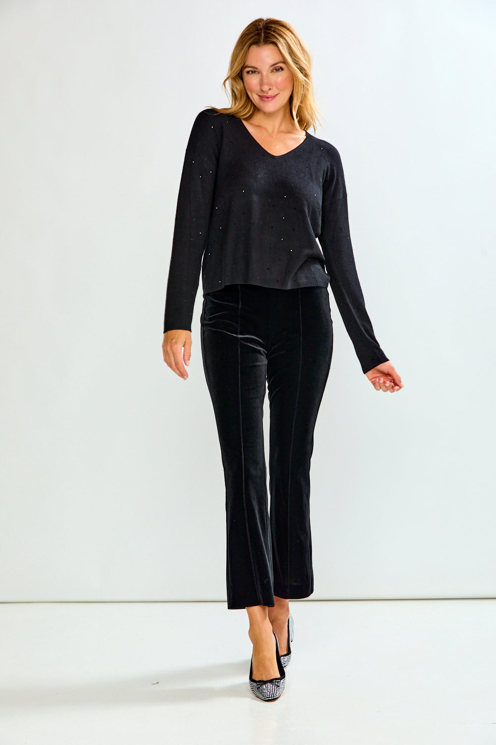 A woman modeling a black long-sleeve V-neck top with subtle crystal embellishments, paired with black pants, against a light gray studio background.