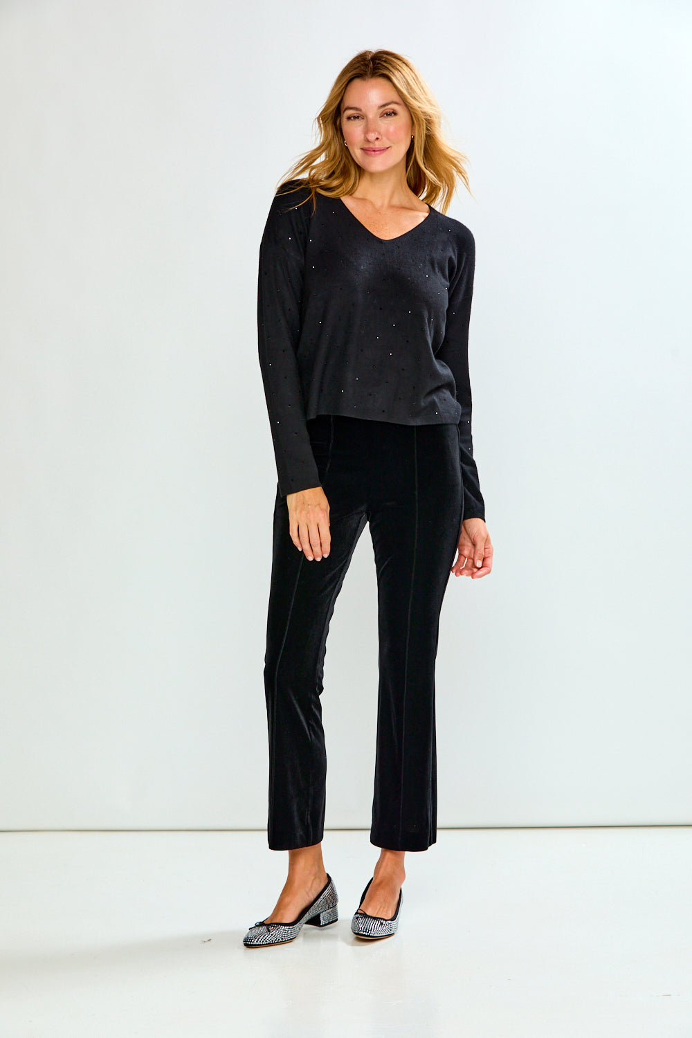 A woman modeling a black long-sleeve knit top with subtle sequin embellishments and black velvet cropped pants, paired with gray pointed-toe flats, against a plain white studio background.