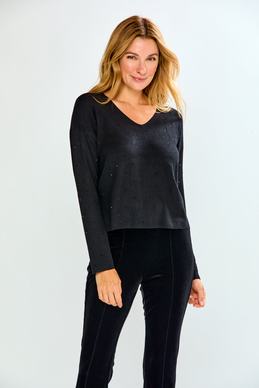 A woman modeling a black long-sleeve V-neck top with subtle crystal embellishments, paired with black pants, against a light gray studio background.