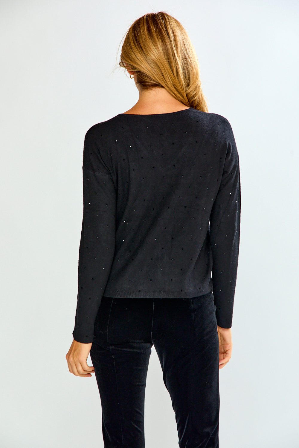A woman modeling a black long-sleeve V-neck top with subtle crystal embellishments, paired with black pants, against a light gray studio background.