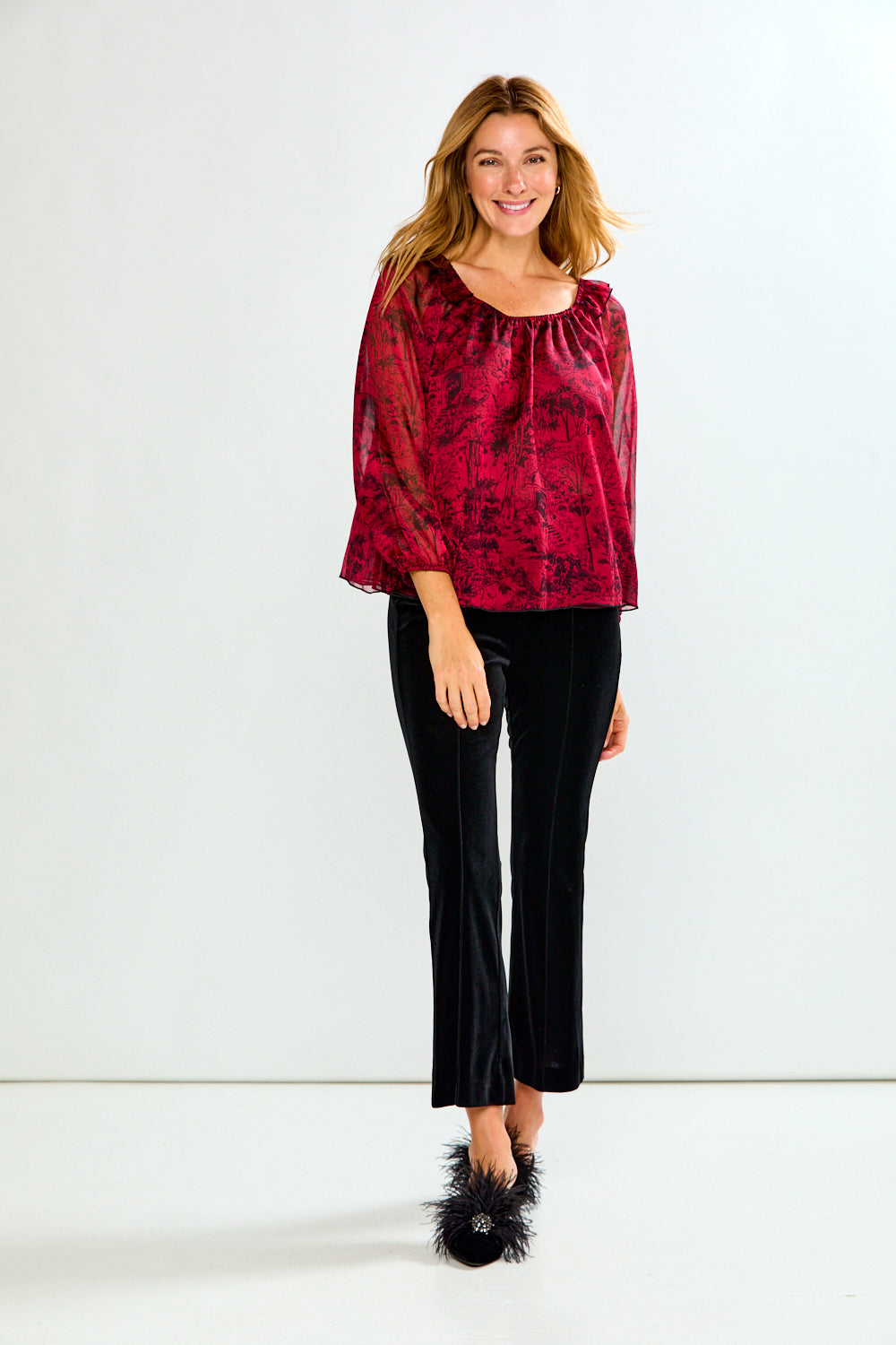 A woman modeling a red and black patterned sheer blouse with flared bell sleeves, paired with black velvet cropped pants and black fuzzy slip-on shoes, against a plain white studio background.