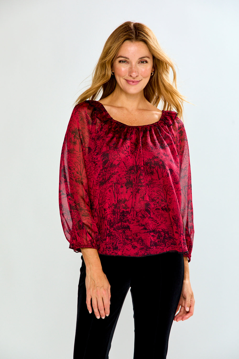 A woman modeling a sheer long-sleeve blouse with a dark red and black botanical pattern, paired with black pants, against a light gray studio background.