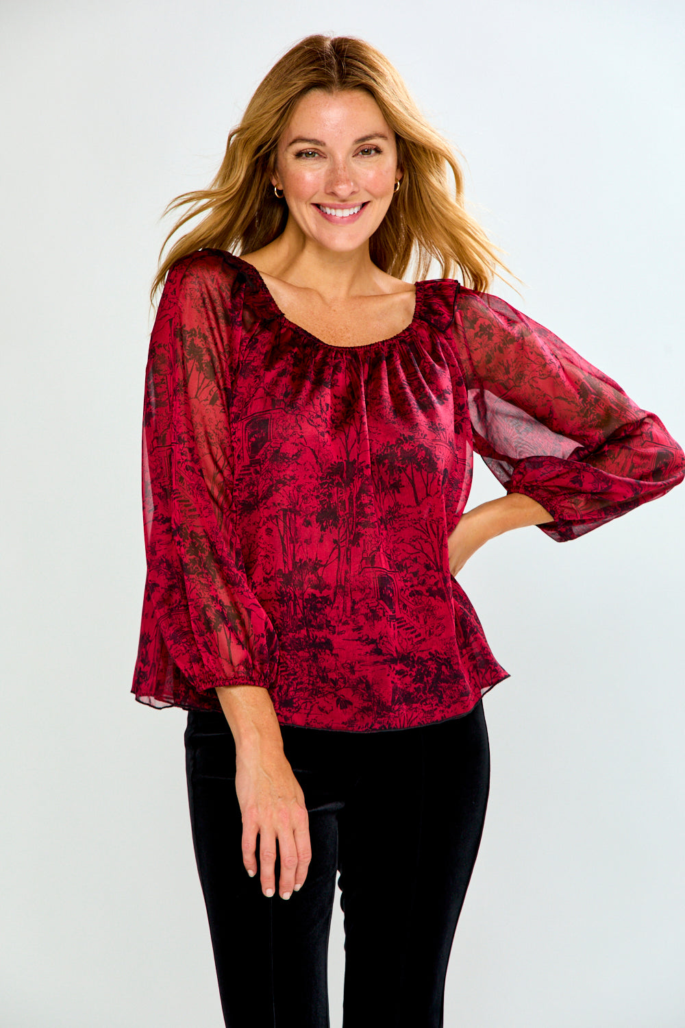 A woman modeling a red and black patterned long-sleeve blouse with a gathered neckline and sheer puff sleeves, paired with black pants, against a light gray studio background.