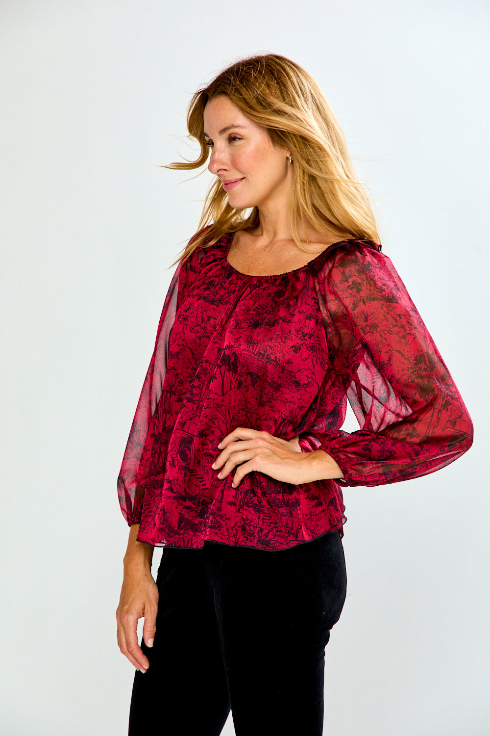 A woman modeling a long-sleeve sheer mesh blouse with a dark red and black floral pattern, paired with black pants, against a light gray studio background.