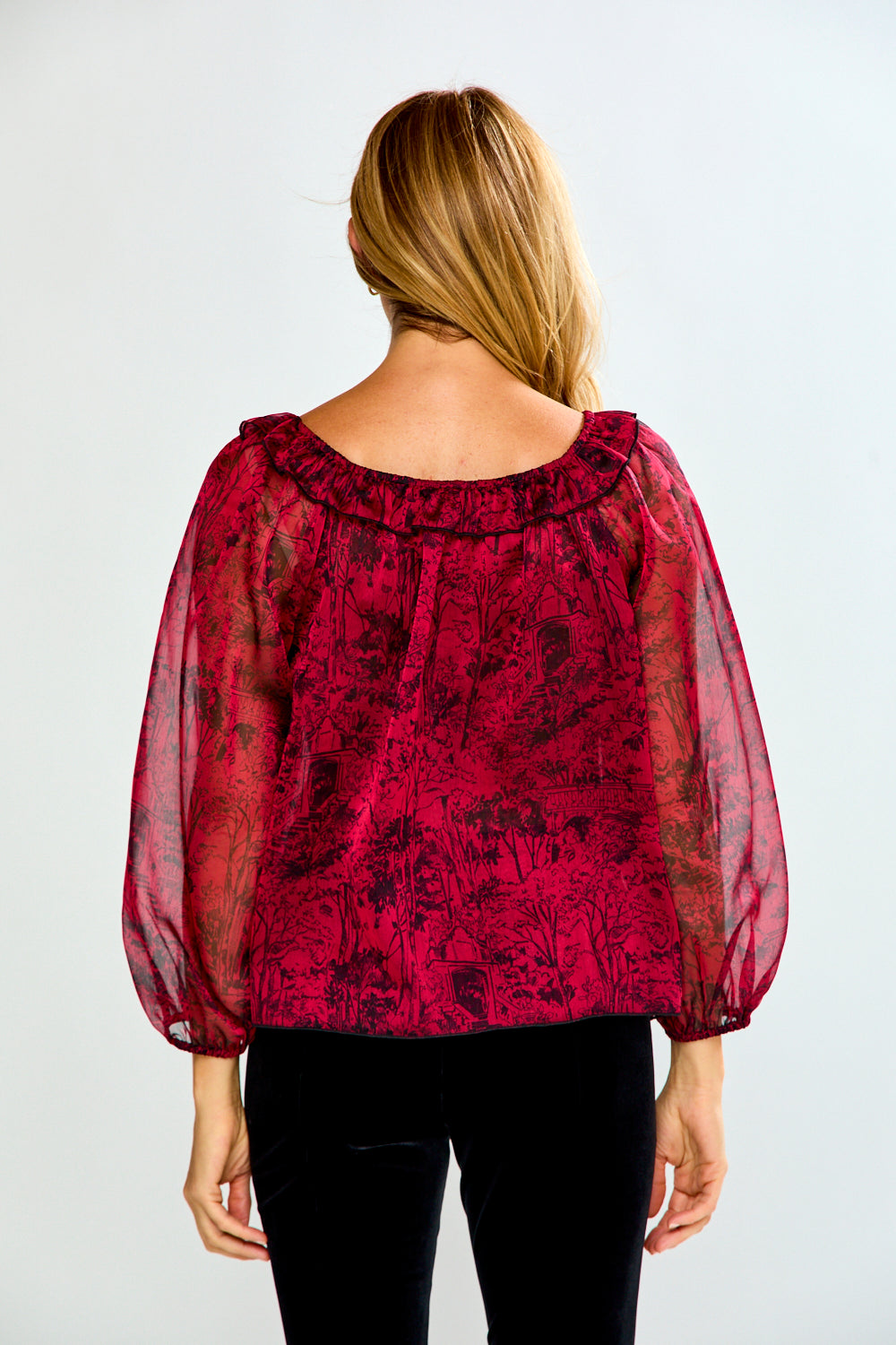 A woman viewed from behind wearing a sheer, long-sleeved blouse with a dark red and black floral toile pattern, paired with black pants, against a light gray background.