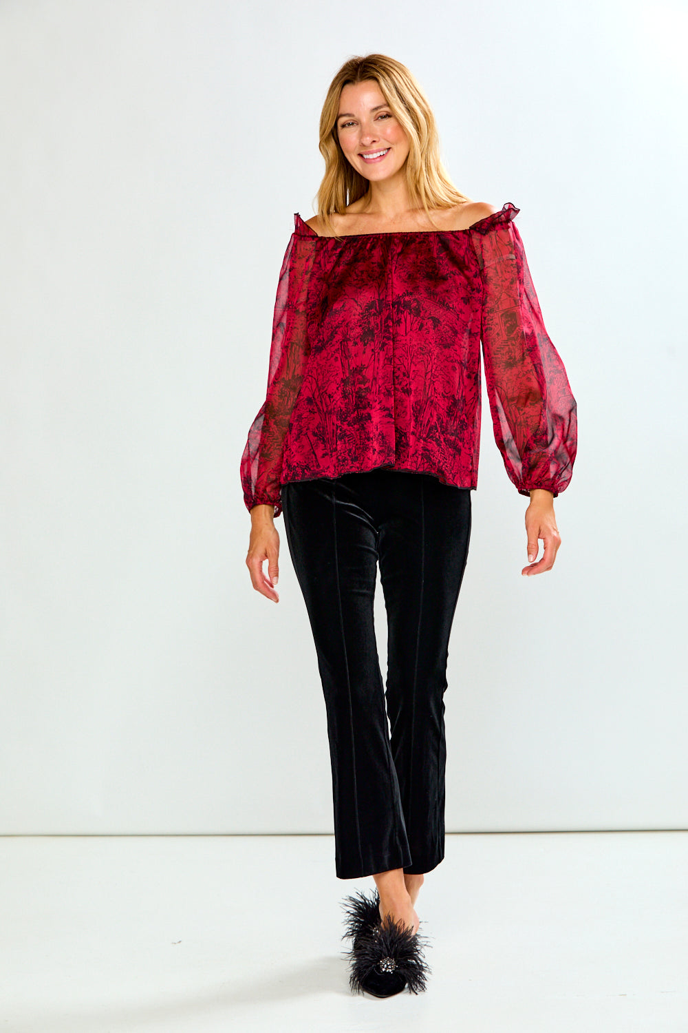 A woman modeling an off-the-shoulder long-sleeve top in a deep red and black damask pattern, paired with black velvet pants and black feathered slippers, against a plain white studio background.