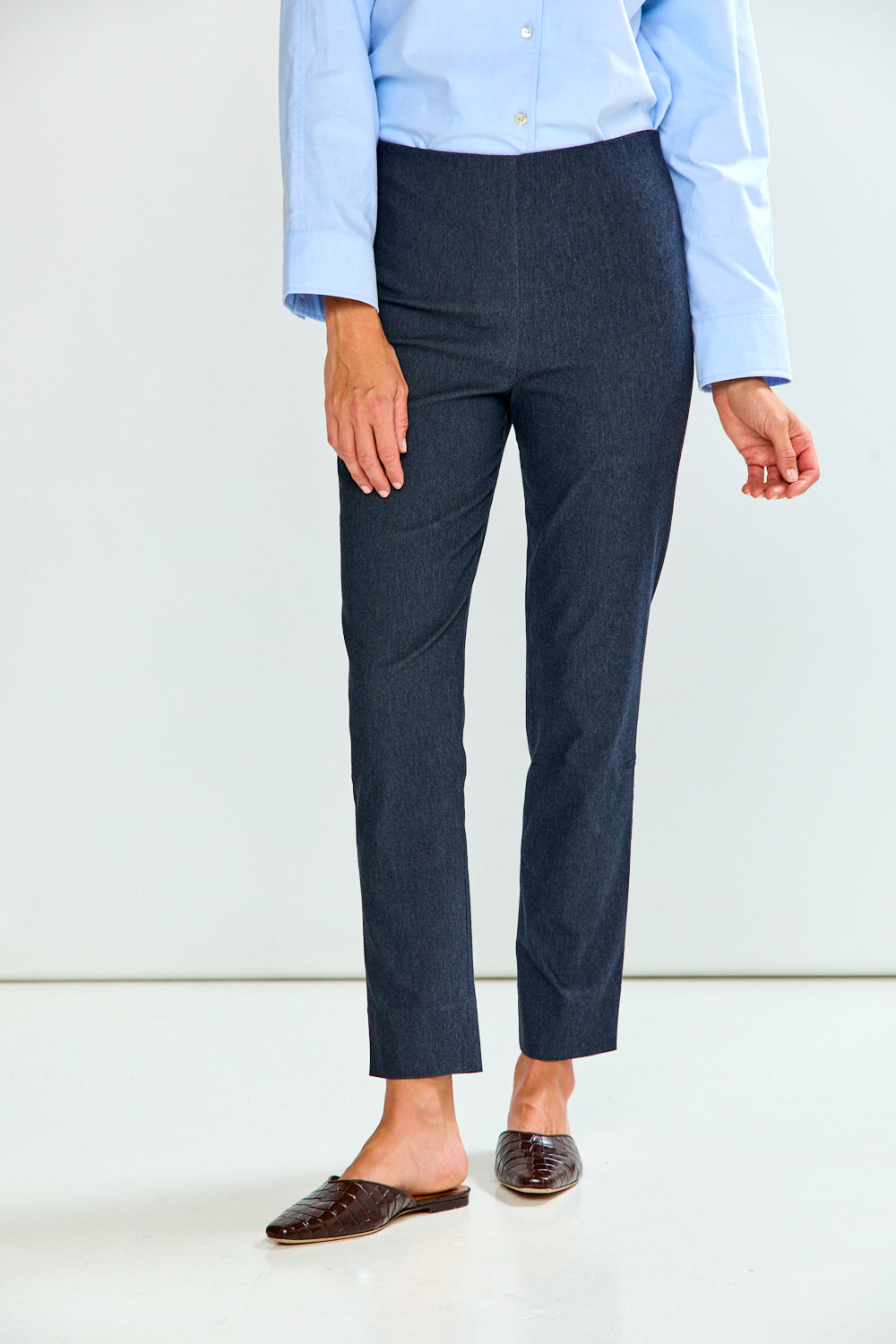 A person wearing denim cropped trousers with a light blue button-up shirt and brown mule flats, standing against a plain light gray studio background.