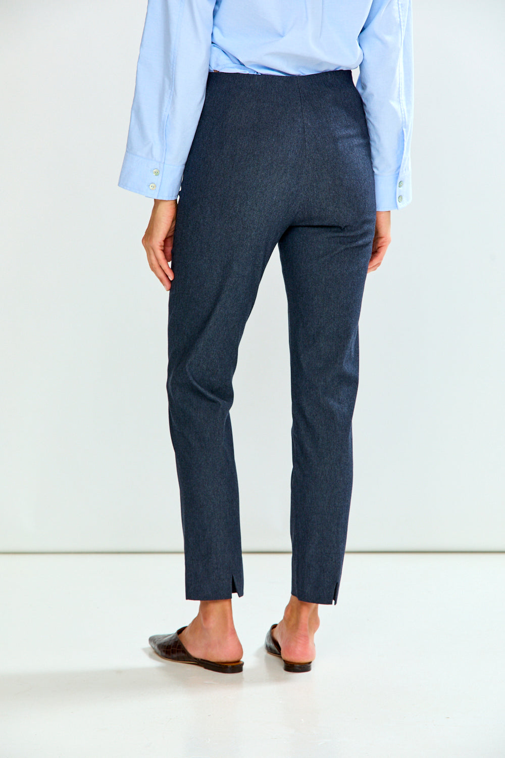 A person wearing denim cropped trousers with a light blue button-up shirt and brown mule flats, standing against a plain light gray studio background.