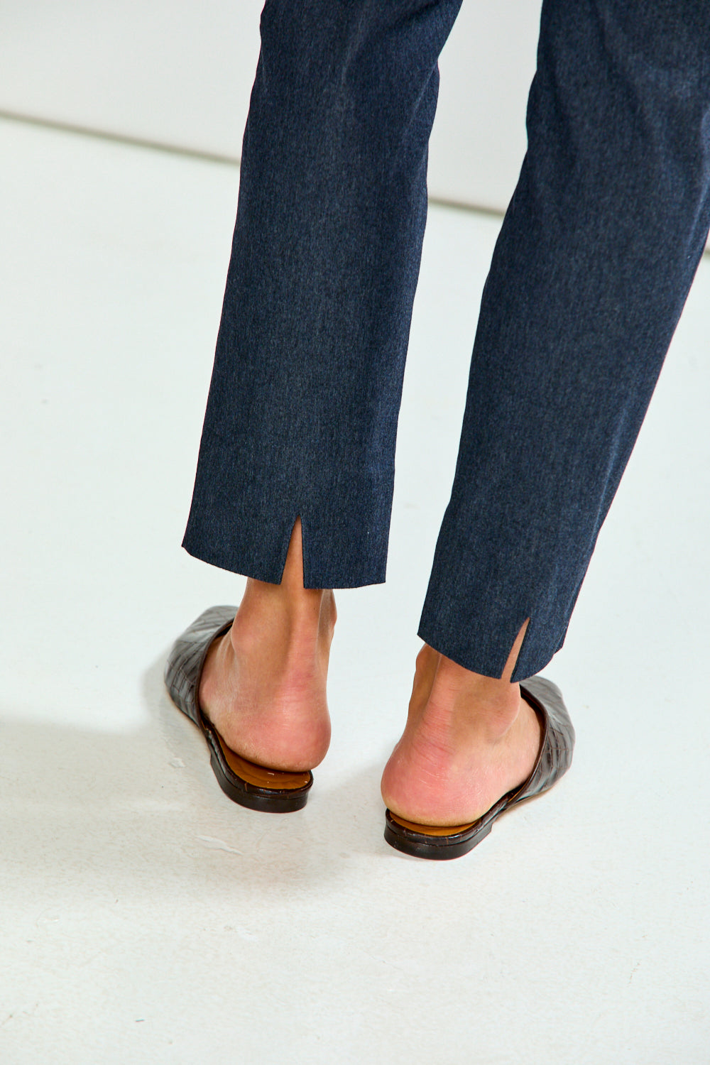 A person wearing denim cropped trousers with a light blue button-up shirt and brown mule flats, standing against a plain light gray studio background.