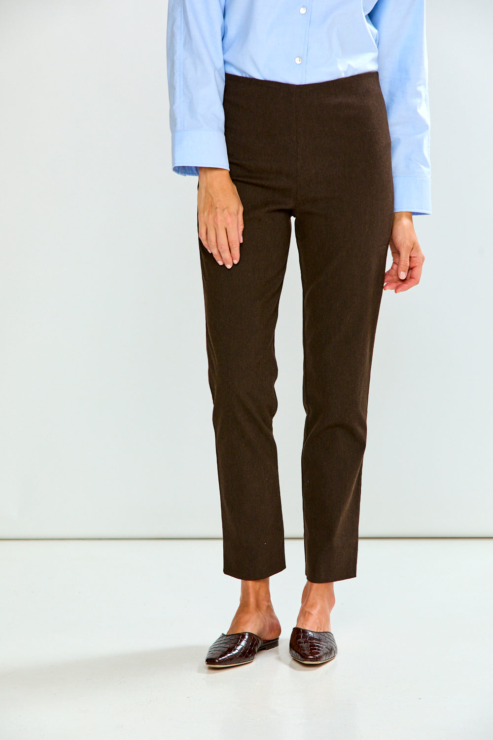 A person wearing dark brown cropped trousers, a light blue button-up shirt, and dark brown mule shoes, standing against a plain light gray studio background.