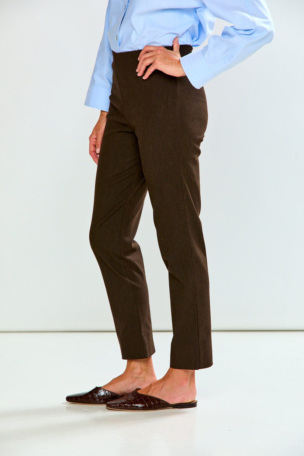 A person wearing dark brown tailored trousers, a light blue long-sleeve shirt, and dark brown loafers, standing against a plain light gray studio background.
