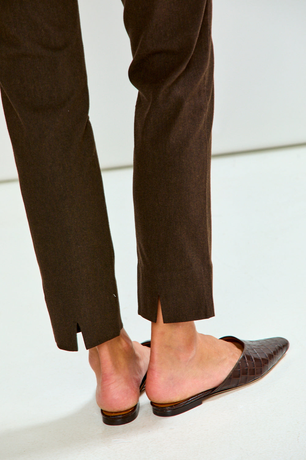 A close-up of a person's feet wearing dark brown pointed-toe mule flats with a crocodile-embossed texture, paired with dark brown trousers.