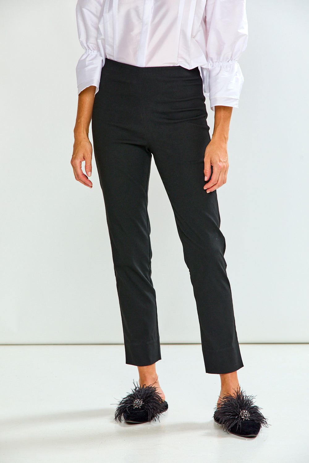 A model wearing black cropped trousers, a white long-sleeve blouse, and black feathered mule flats, shown from the waist down against a plain white studio background.