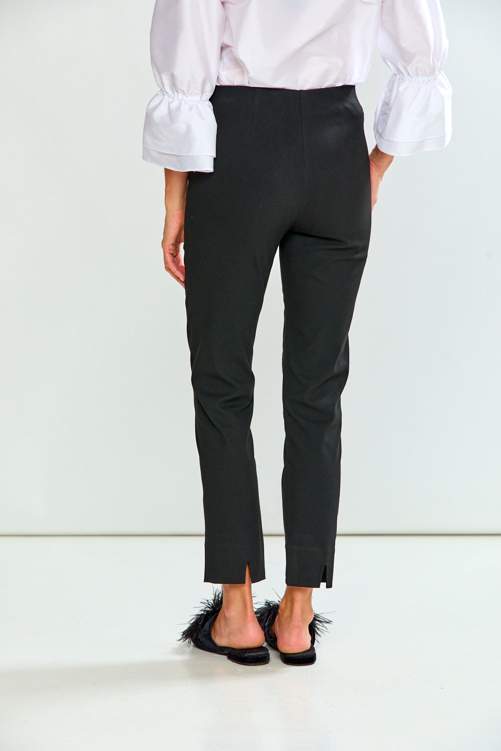 A person shown from the back wearing black cropped trousers, a white blouse with ruffled sleeves, and black feathered mule sandals against a clean white studio background.