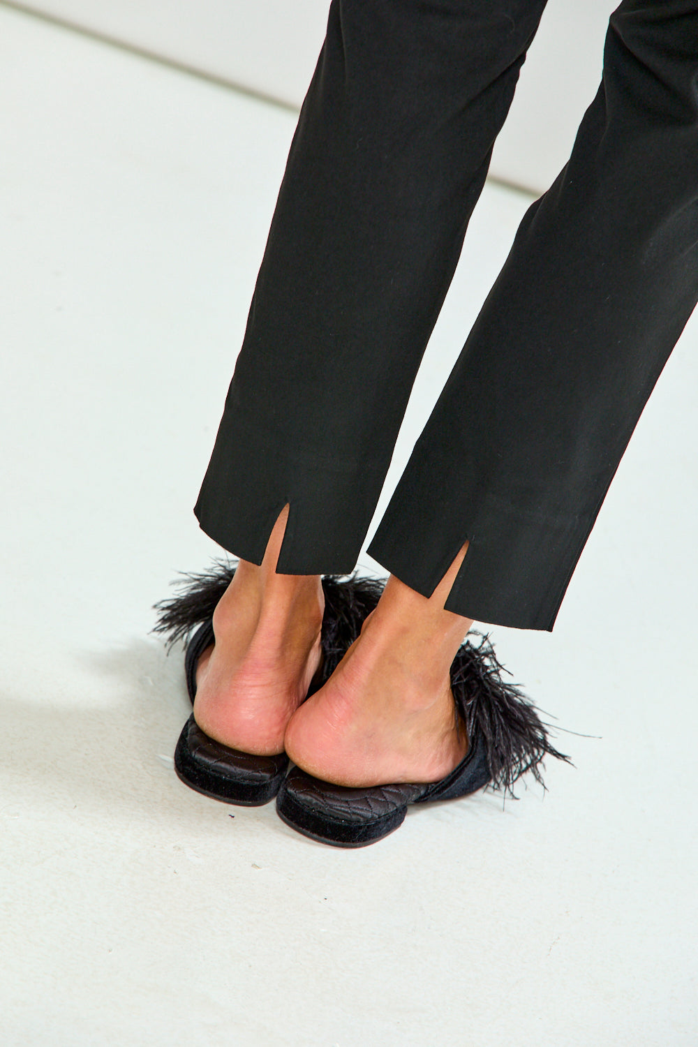 Black flat mule sandals with black feather trim, shown on a model's feet wearing black trousers against a clean white background.