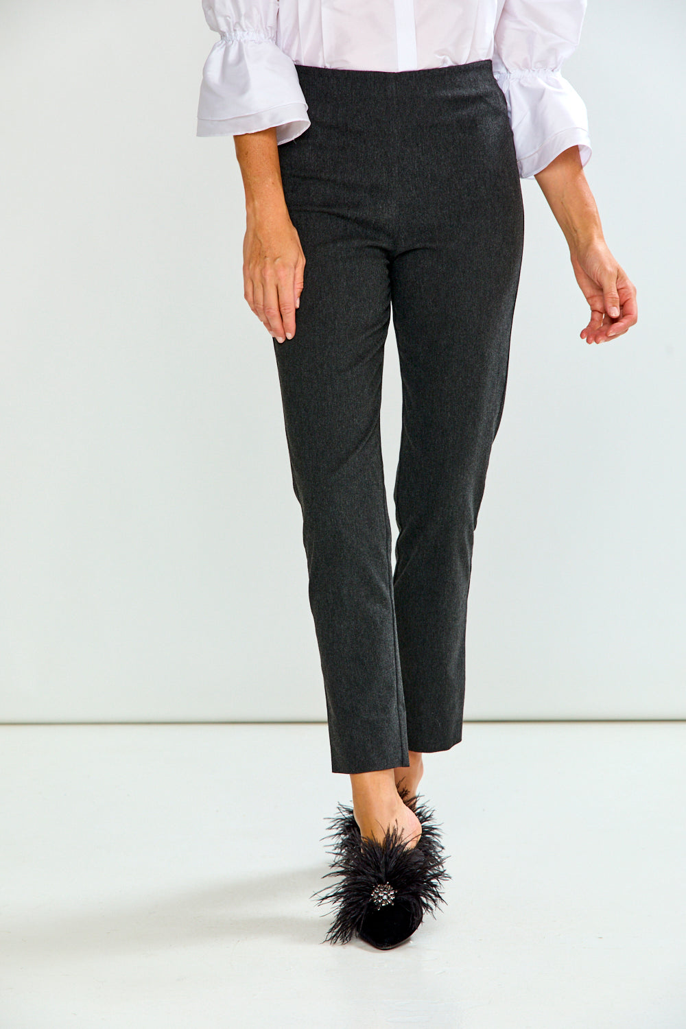 A model is shown from the waist down wearing dark charcoal gray cropped trousers, a white blouse with ruffled sleeves, and black feathered mule shoes against a clean white studio background.