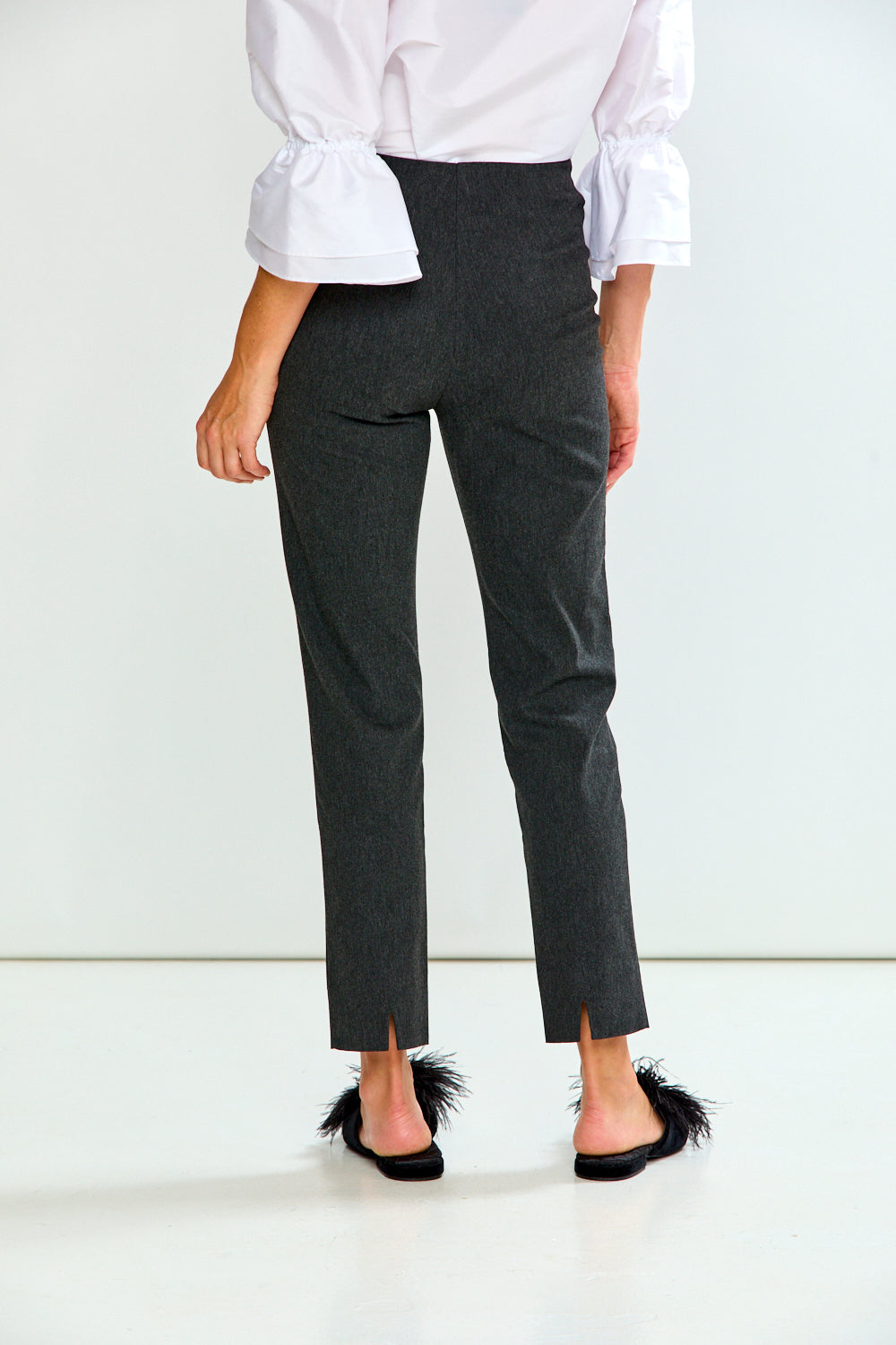 A model from the back wearing dark charcoal gray cropped trousers, a white blouse with ruffled sleeves, and black mule sandals with feather trim, set against a clean white studio background.