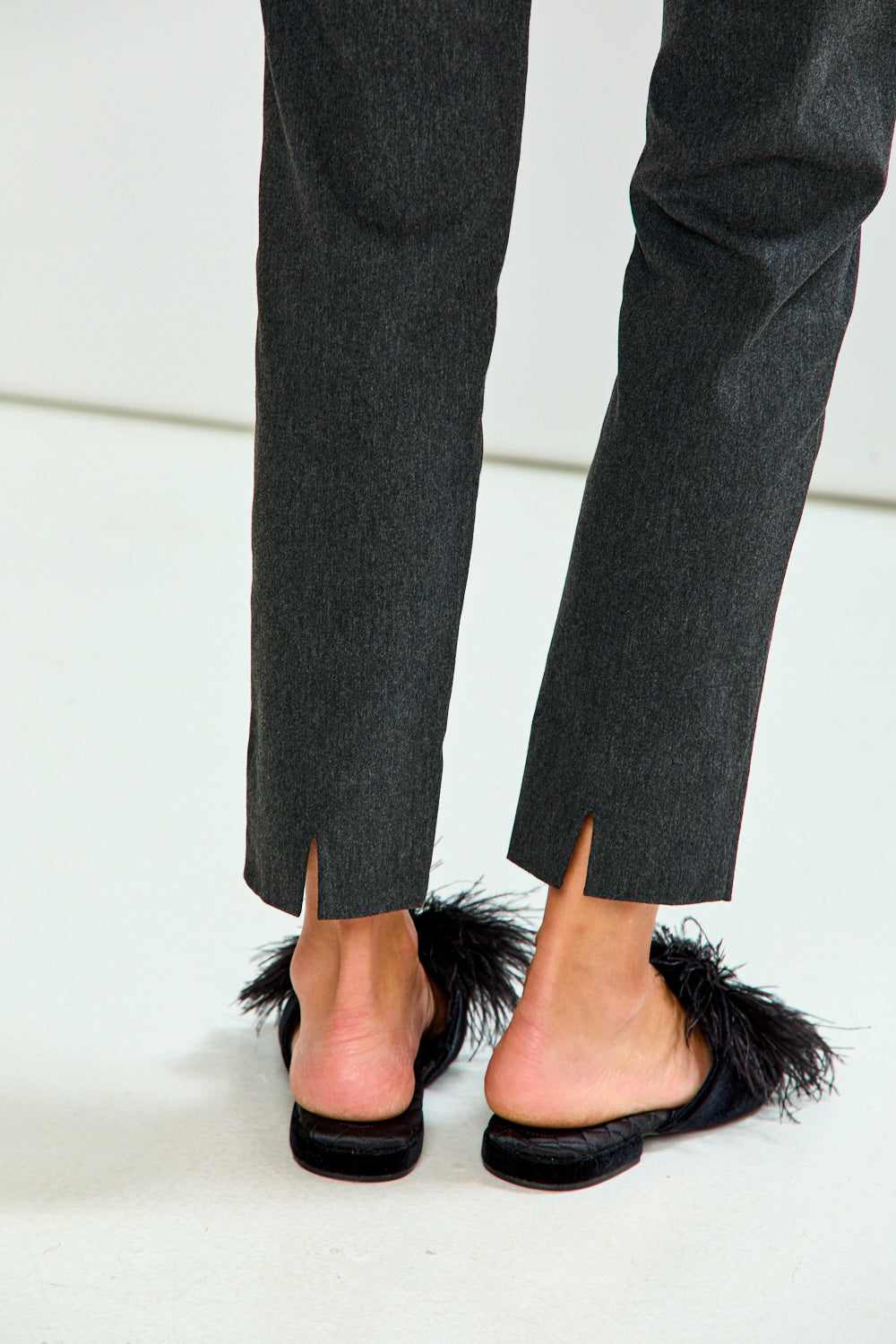 Close-up rear view of a person wearing dark charcoal gray trousers and black mule sandals with feather trim, standing against a clean white studio background.