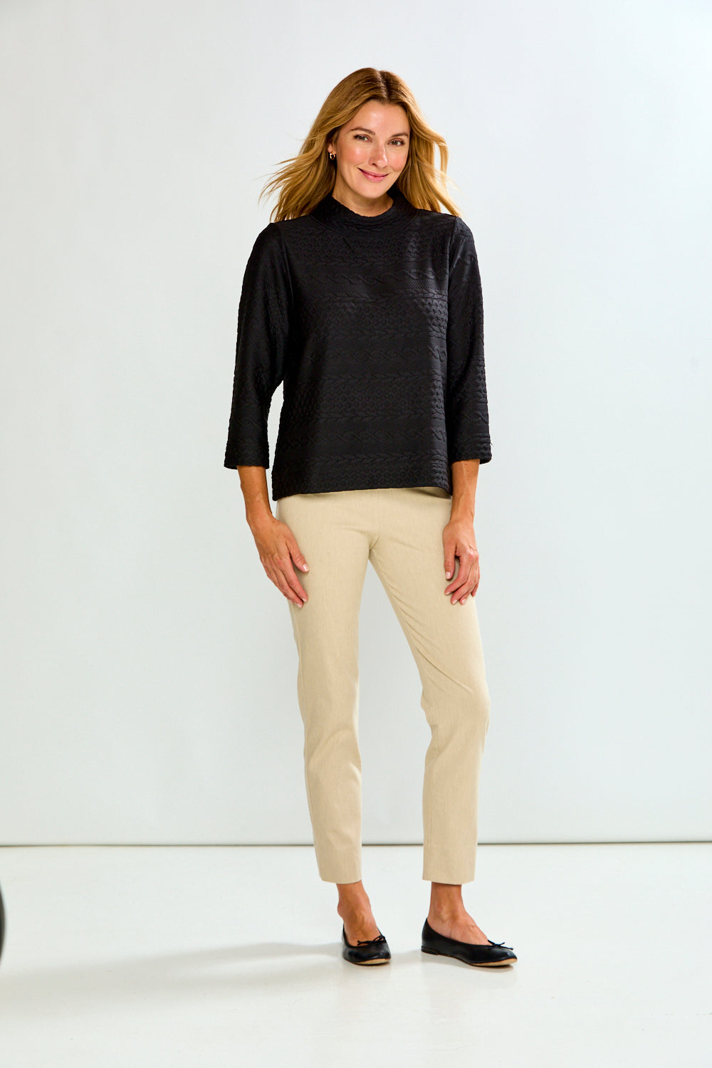 A model wearing sand cropped trousers, a black long-sleeve top, and black flat shoes, standing against a plain white studio background.