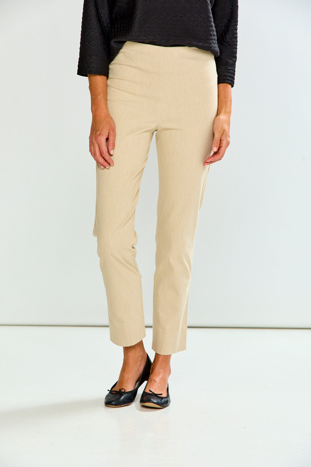 A model wearing sand cropped trousers, a black long-sleeve top, and black flat shoes, standing against a plain white studio background.