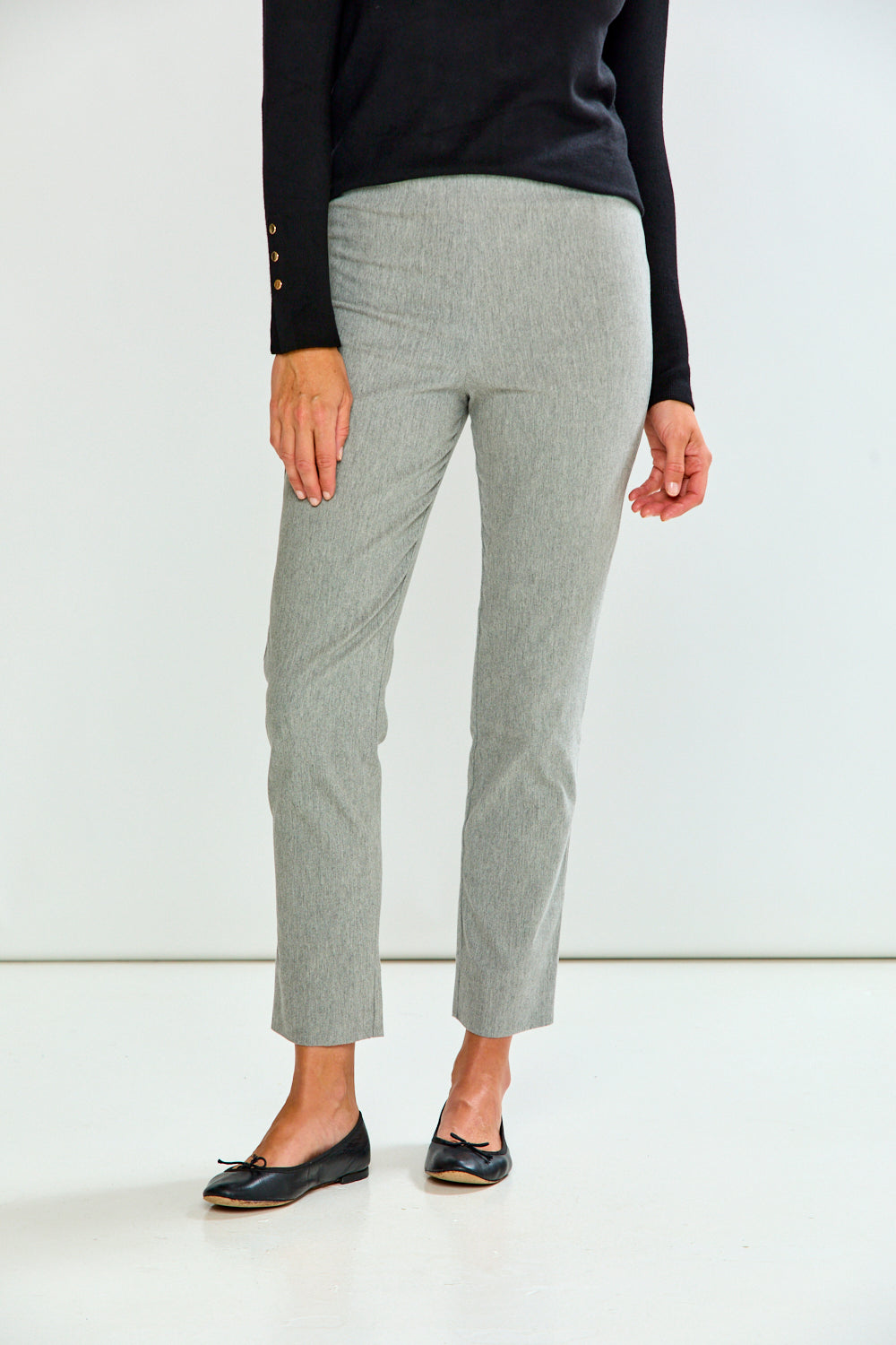 A person wearing light gray cropped trousers, a black long-sleeve top, and black flats, standing against a plain white studio background.