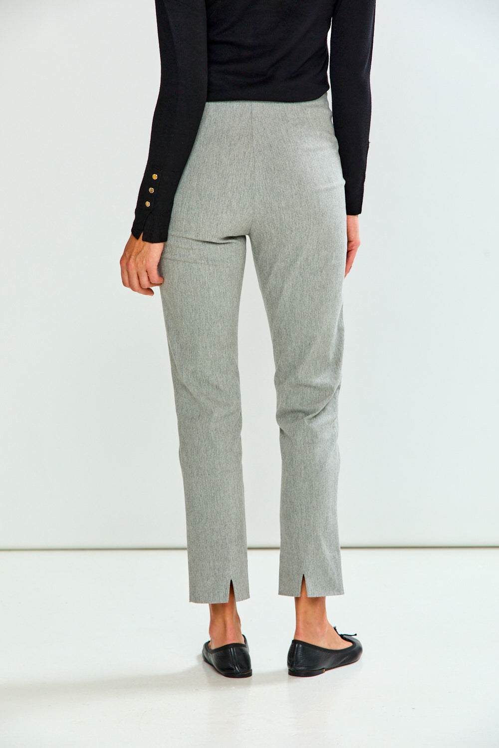 A person shown from the back wearing light gray cropped trousers with side slits, a black long-sleeve top with gold buttons, and black flats, against a clean white studio background.