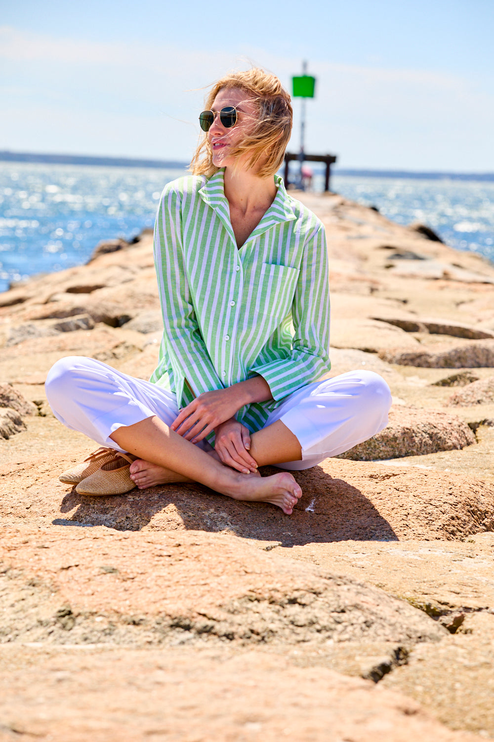 Woman sitting on a rocky beach wearing a green and white striped shirt and white pants.