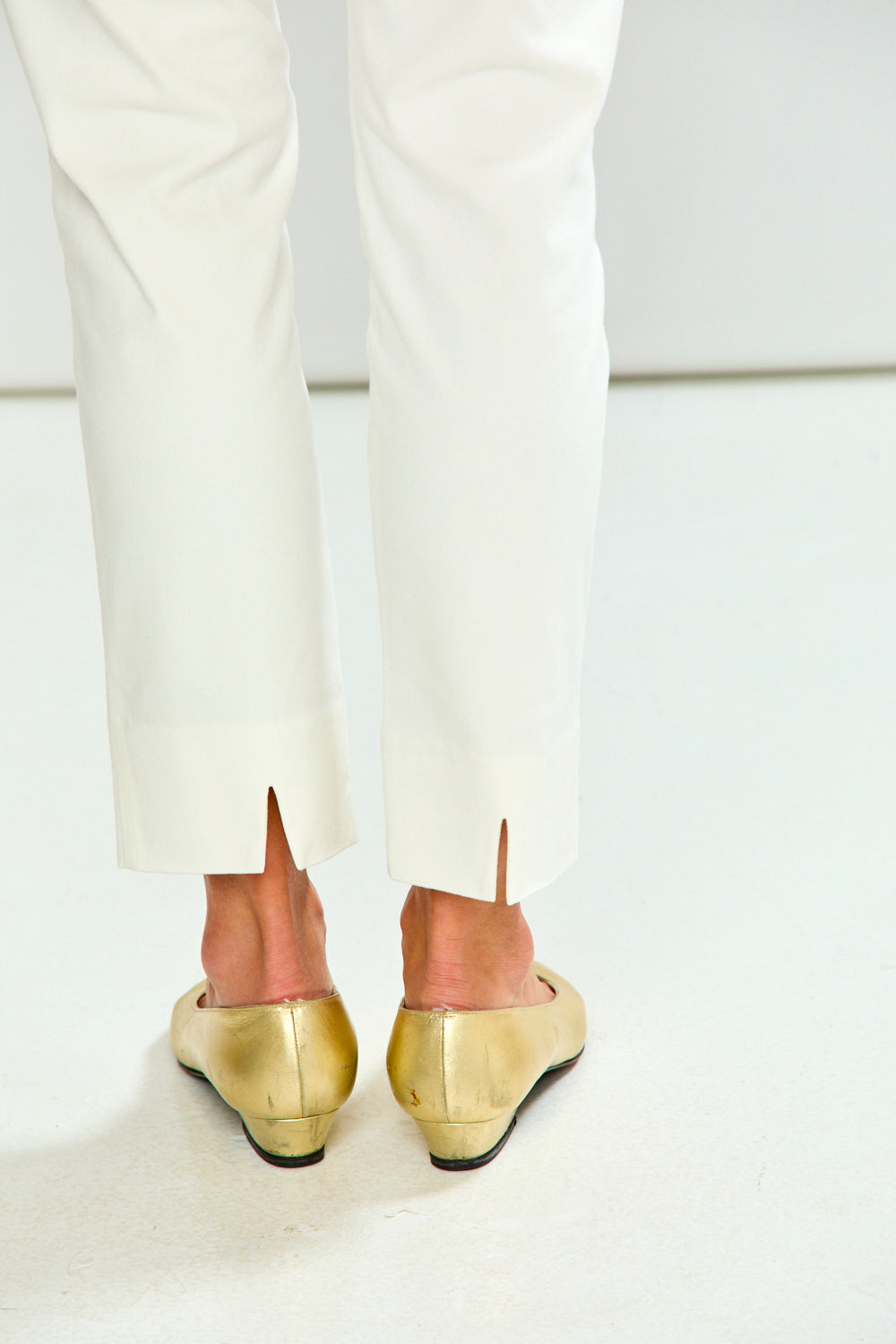 A close-up rear view of a person wearing white trousers and shiny metallic gold wedge shoes against a clean white studio background.