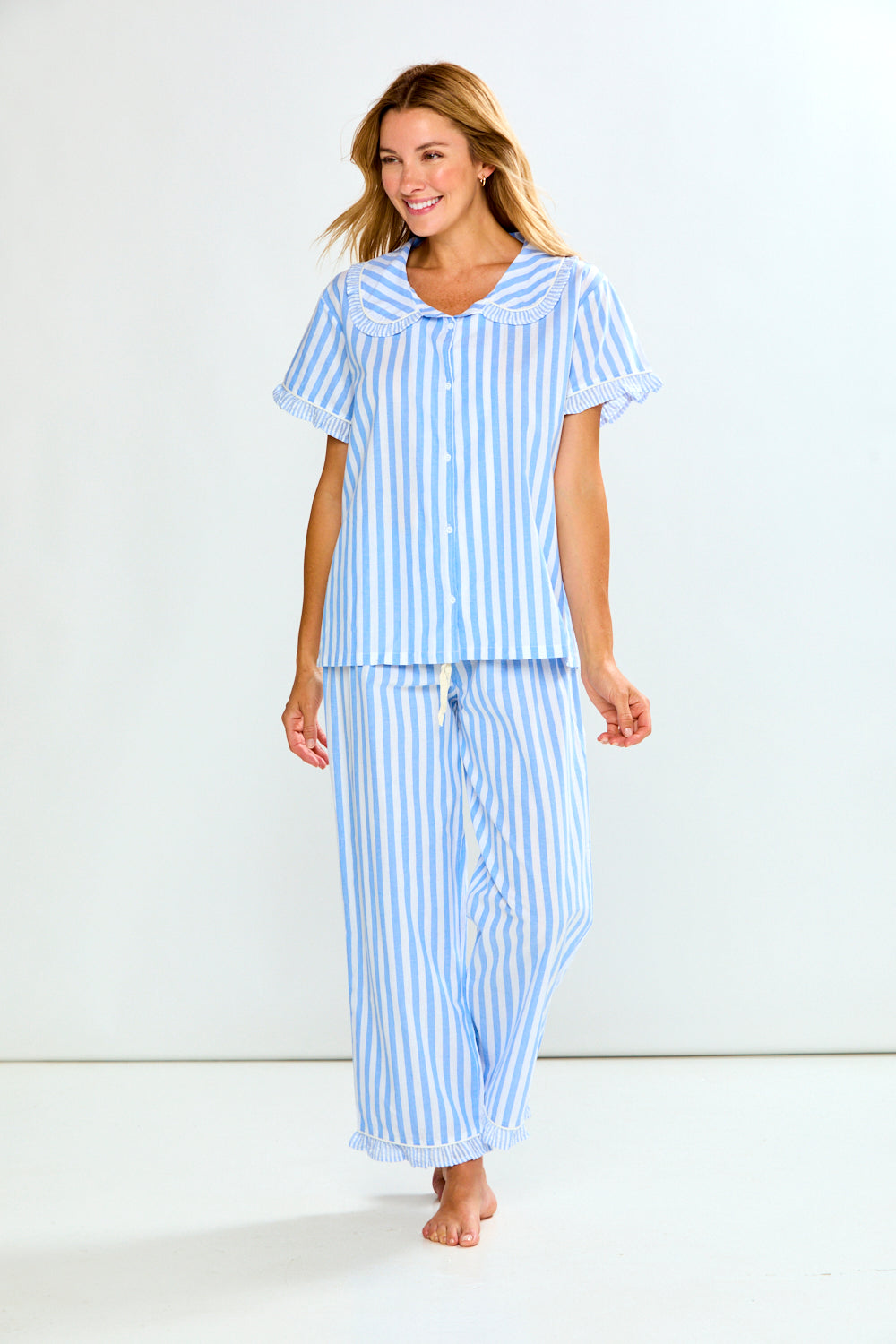 A woman modeling a light blue and white vertically striped pajama set, consisting of a short-sleeve button-up top with ruffled trim and matching long pants, against a plain white studio background.