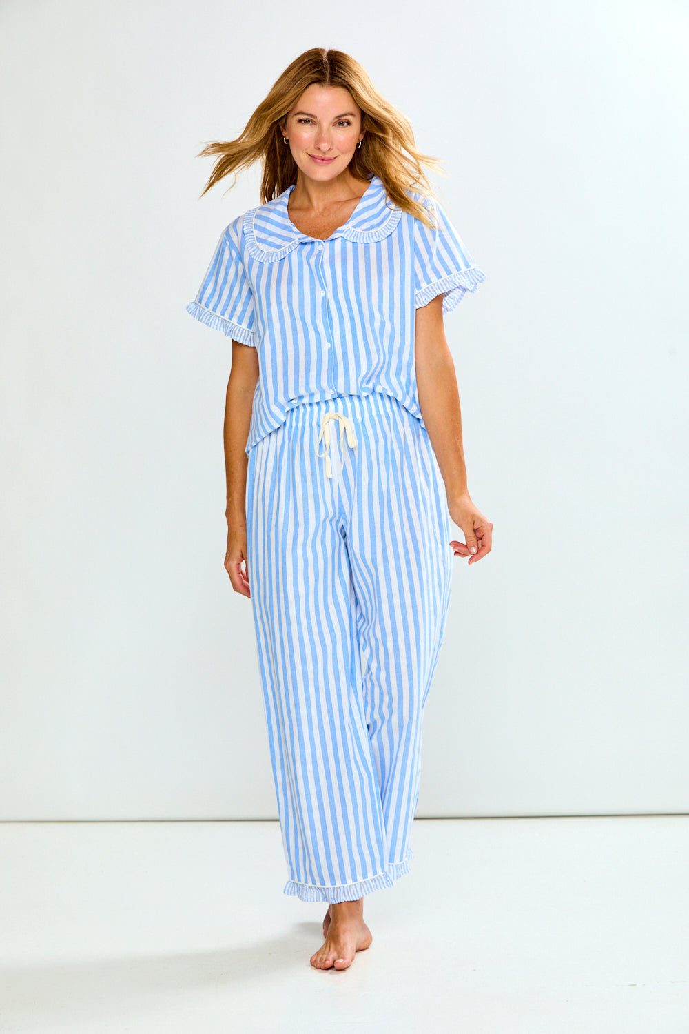 A woman modeling a light blue and white vertically striped pajama set with a short-sleeve button-front top and matching drawstring pants, photographed against a plain white studio background.