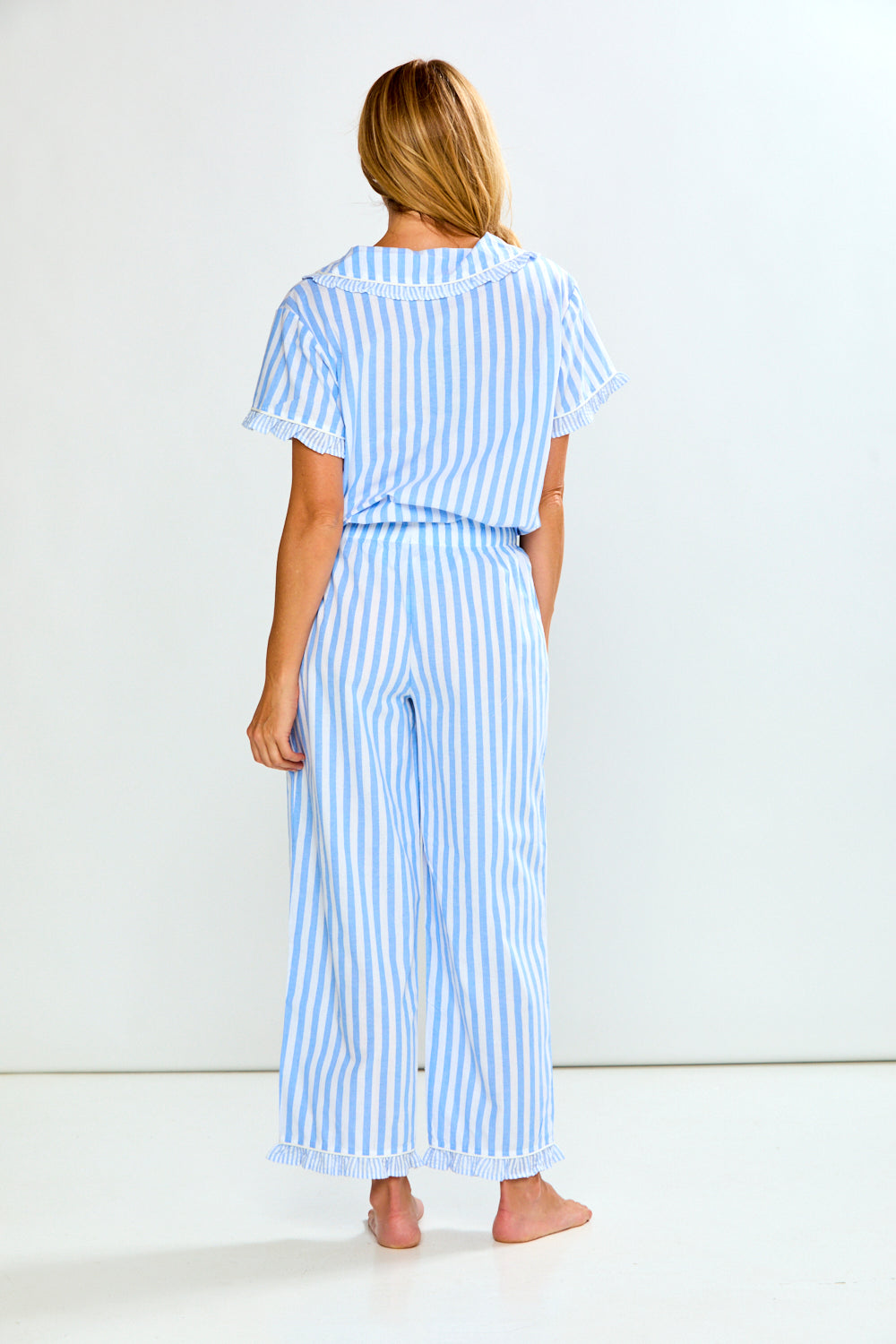 A woman viewed from behind wearing a light blue and white vertically striped two-piece pajama set with a short-sleeve top and wide-leg pants, standing barefoot against a plain white studio background.