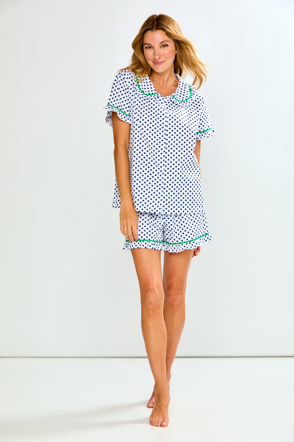 A woman modeling a two-piece pajama set consisting of a short-sleeve top and matching shorts, featuring a white base with a blue polka dot pattern and green trim details.