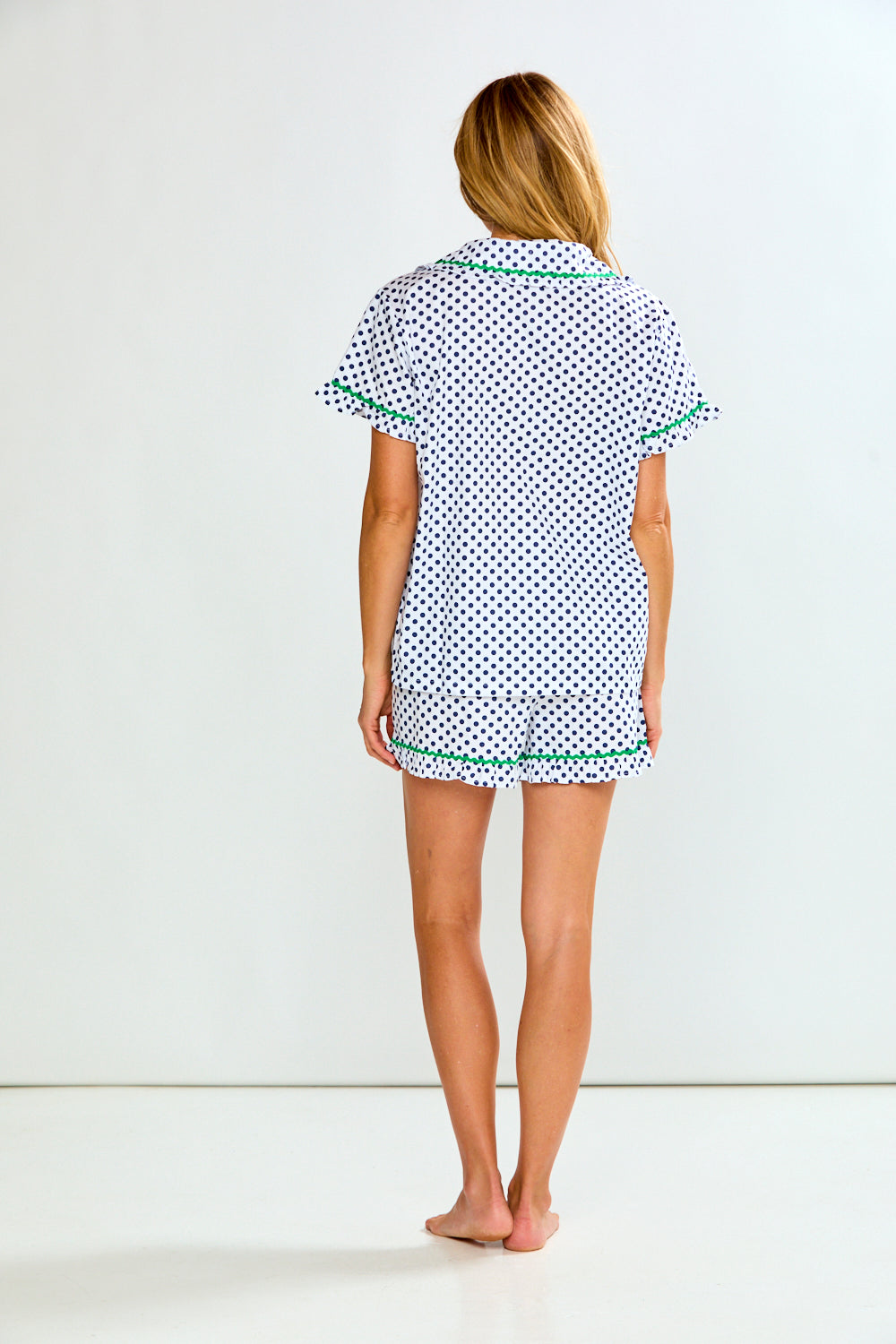 A woman modeling a two-piece pajama set consisting of a short-sleeve top and matching shorts, featuring a white base with a blue polka dot pattern and green trim details.