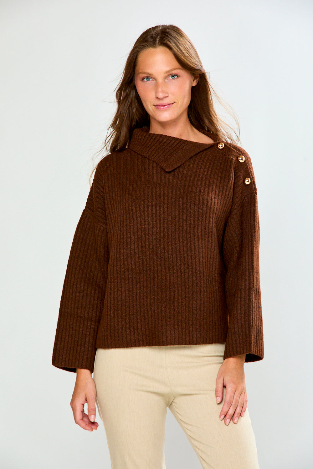 A woman modeling a brown ribbed knit sweater with a wide collar and decorative gold buttons on the shoulder, paired with beige pants, against a light gray studio background.