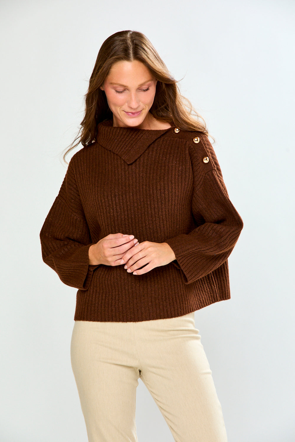 A woman wearing a brown ribbed knit sweater with a wide collar and decorative buttons on the shoulders, paired with beige pants, against a light gray studio background.