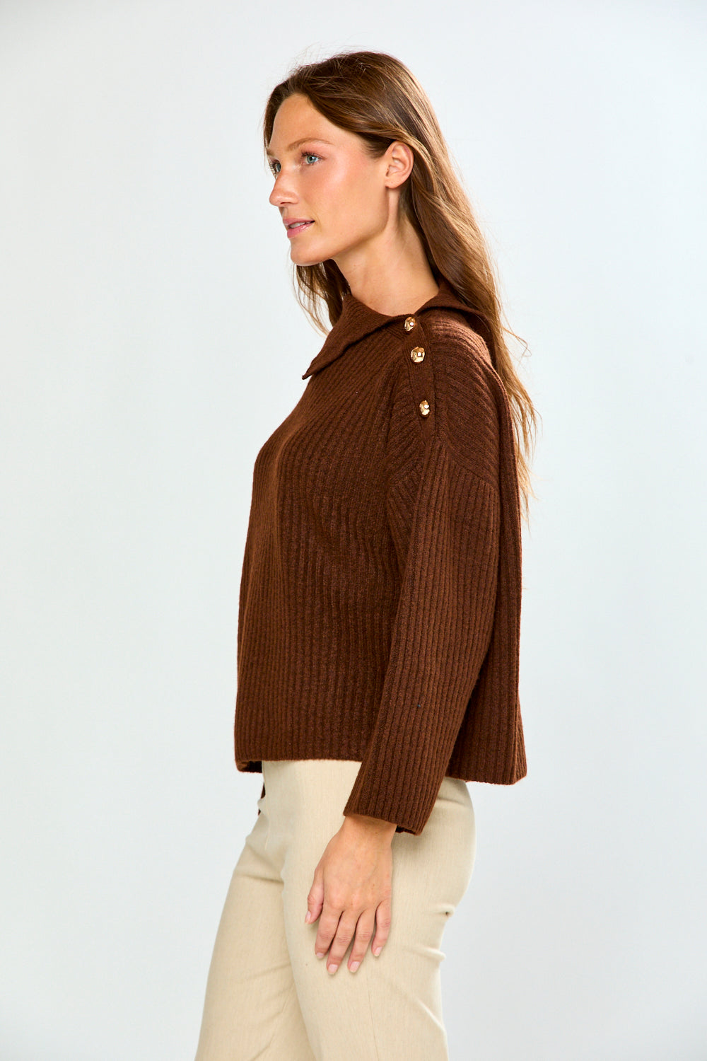 A woman in side profile wearing a brown ribbed knit sweater with decorative gold buttons on the shoulder and light beige trousers, photographed against a light gray studio background.