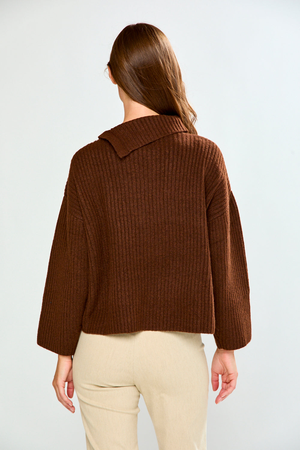A woman shown from the back wearing a brown ribbed knit cropped sweater and light beige pants against a plain light gray studio background.