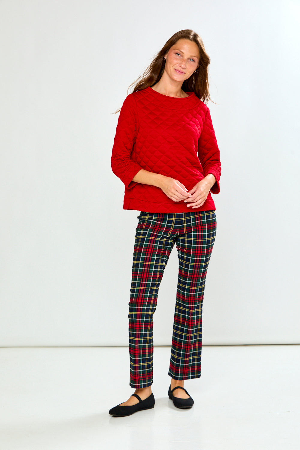 A woman modeling a red long-sleeve quilted knit top with a boat neckline, paired with green and red plaid pants, against a light gray studio background.
