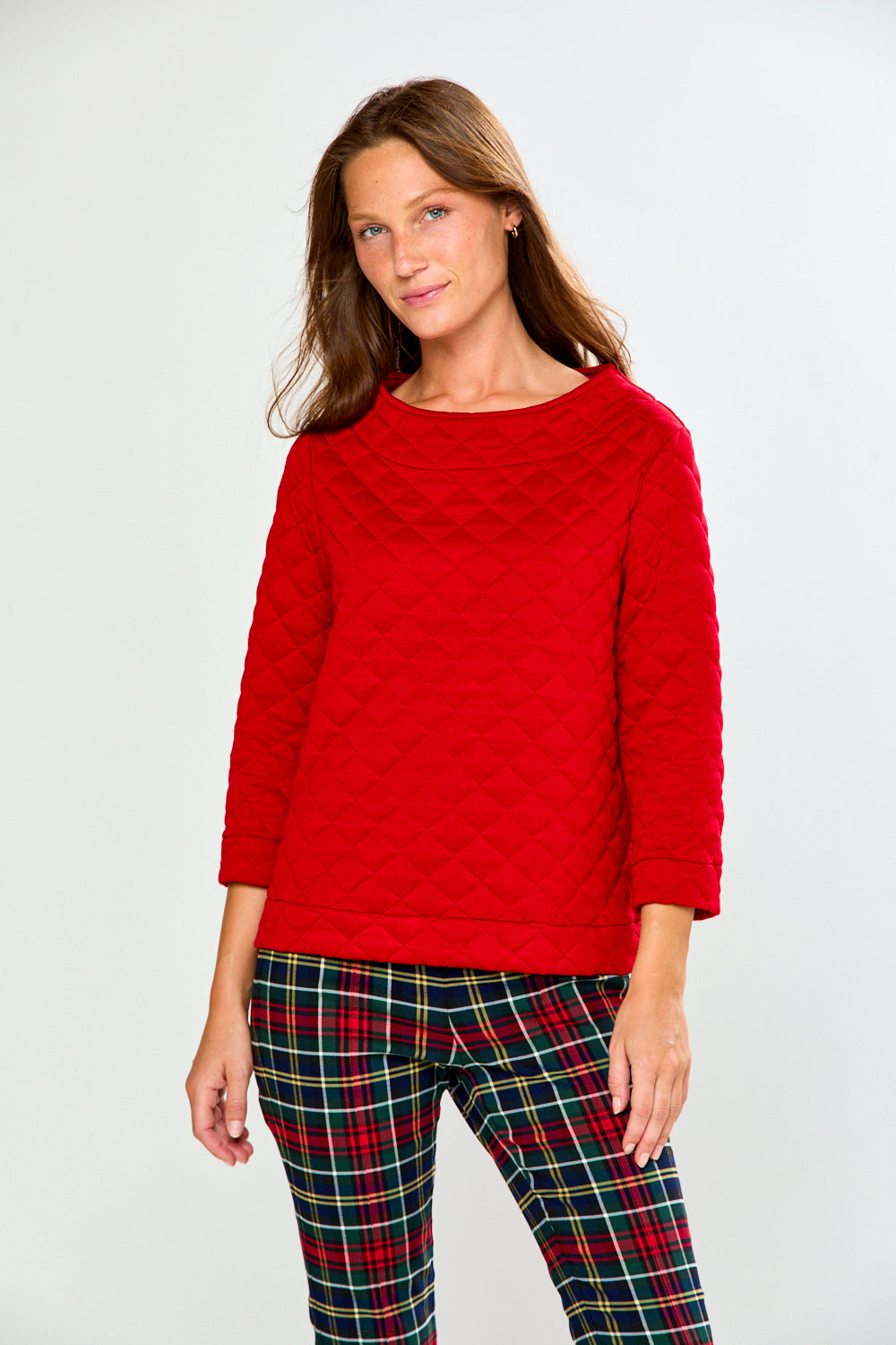 A woman modeling a red long-sleeve quilted knit top with a boat neckline, paired with green and red plaid pants, against a light gray studio background.