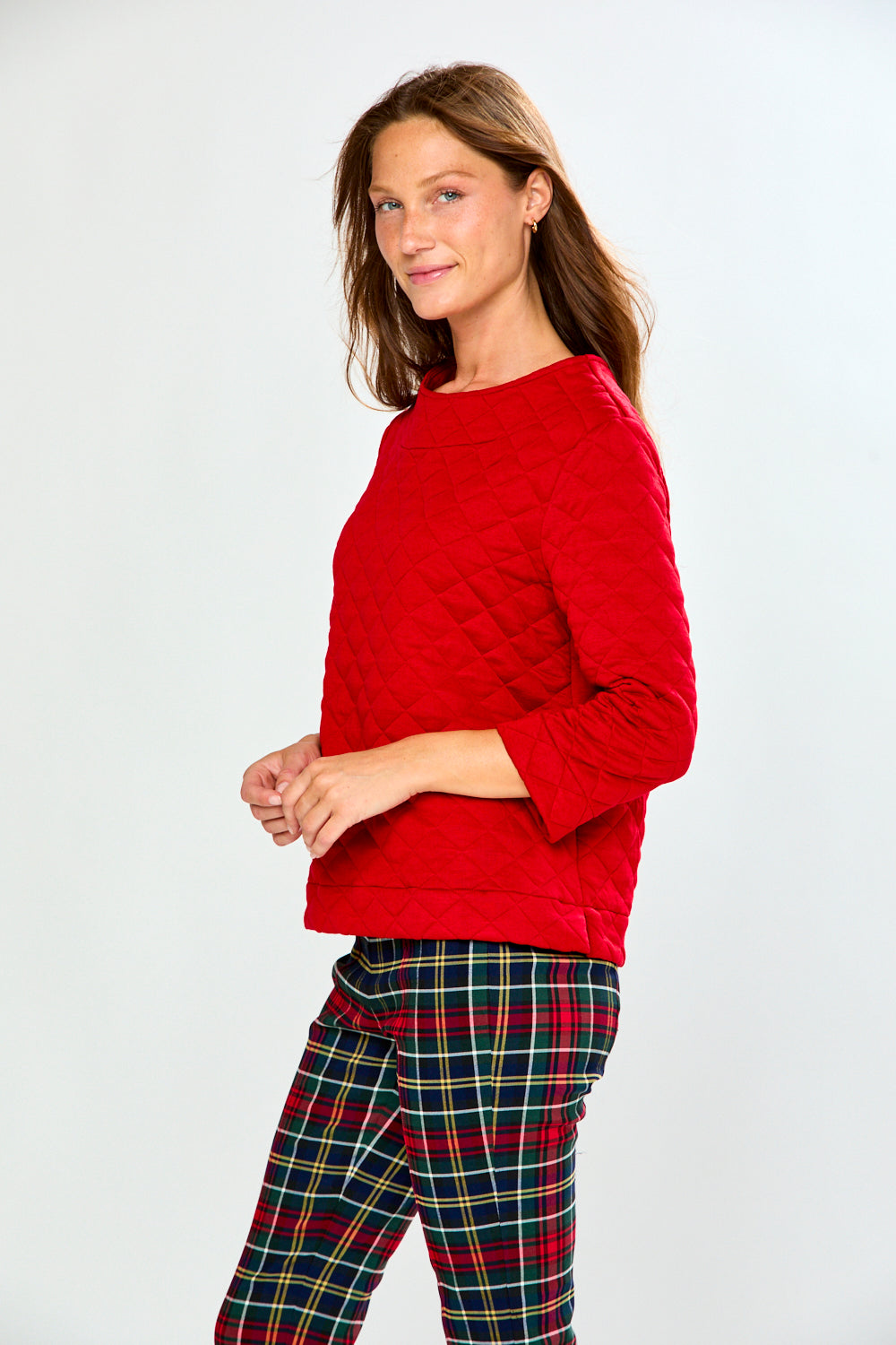 A woman modeling a red long-sleeve quilted knit top with a boat neckline, paired with green and red plaid pants, against a light gray studio background.