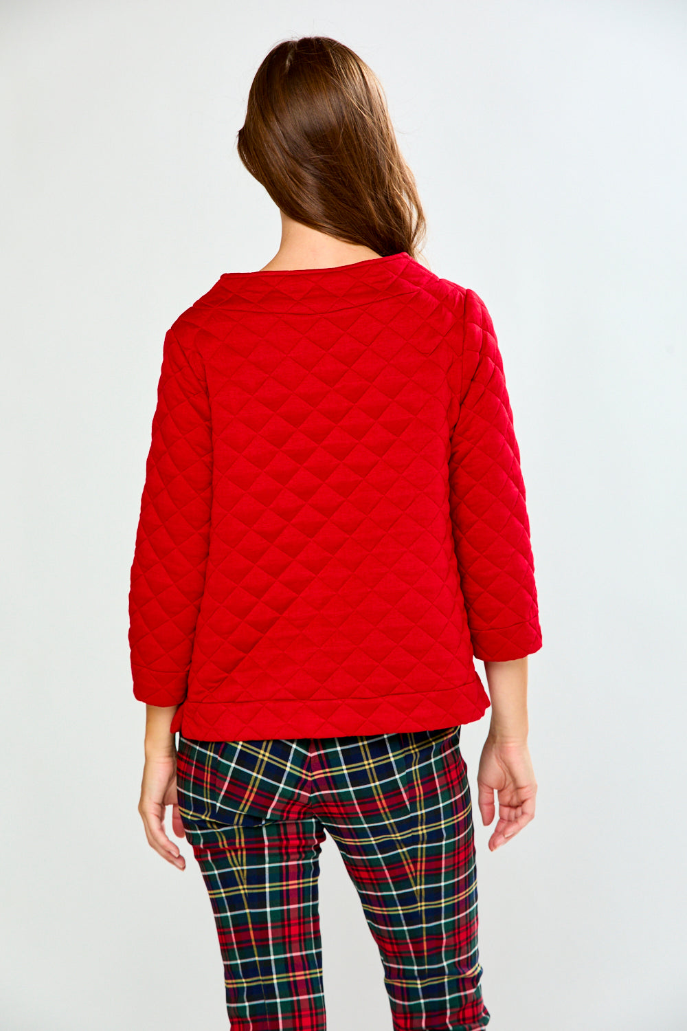 A woman modeling a red long-sleeve quilted knit top with a boat neckline, paired with green and red plaid pants, against a light gray studio background.